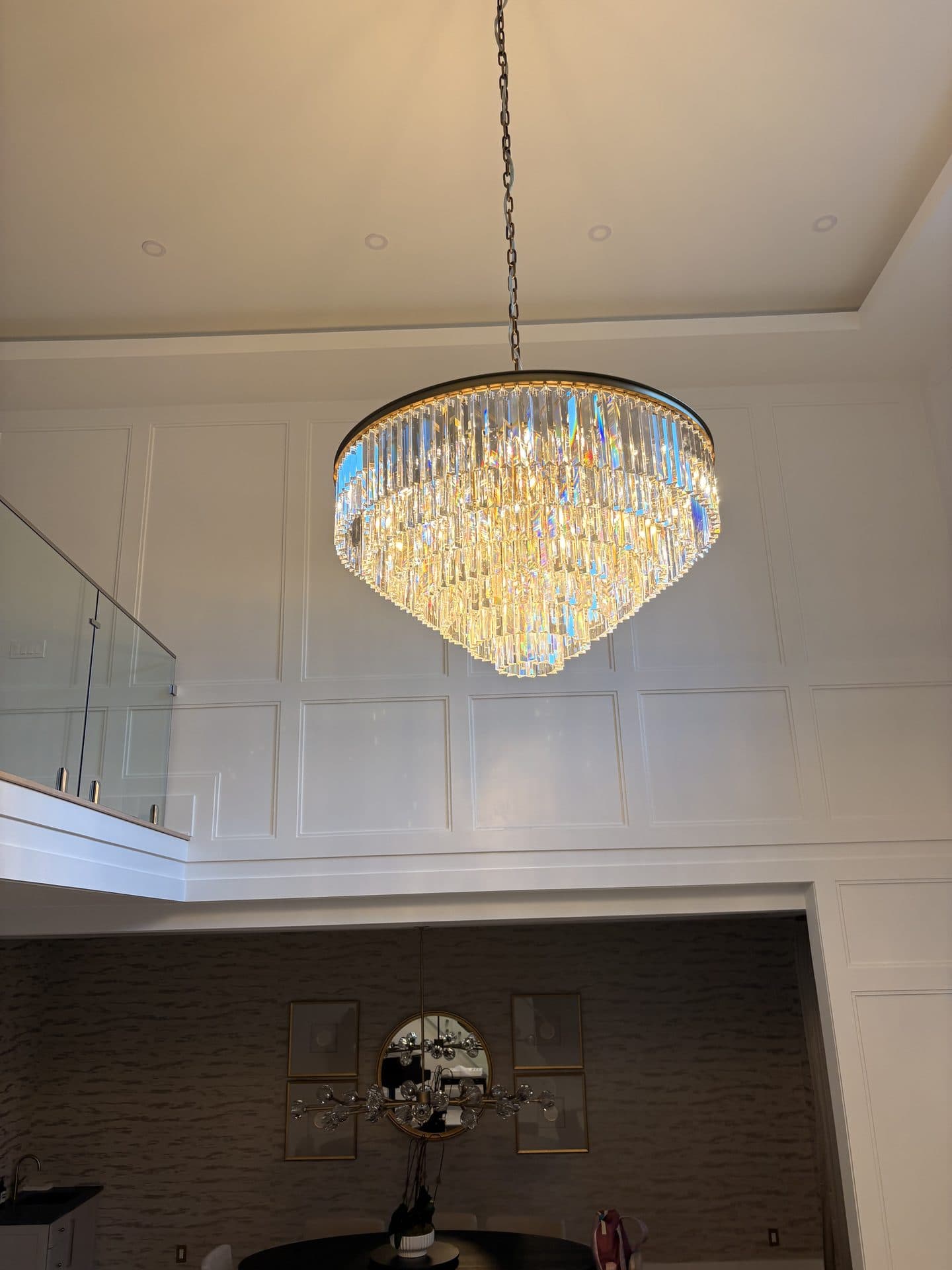 A lit tiered crystal chandelier hanging below a glass-railed balcony in a paneled two-story foyer