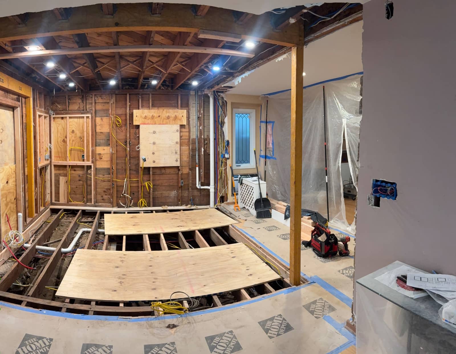 A gutted kitchen mid-renovation with recessed lights on in the open ceiling and wiring run along the studs