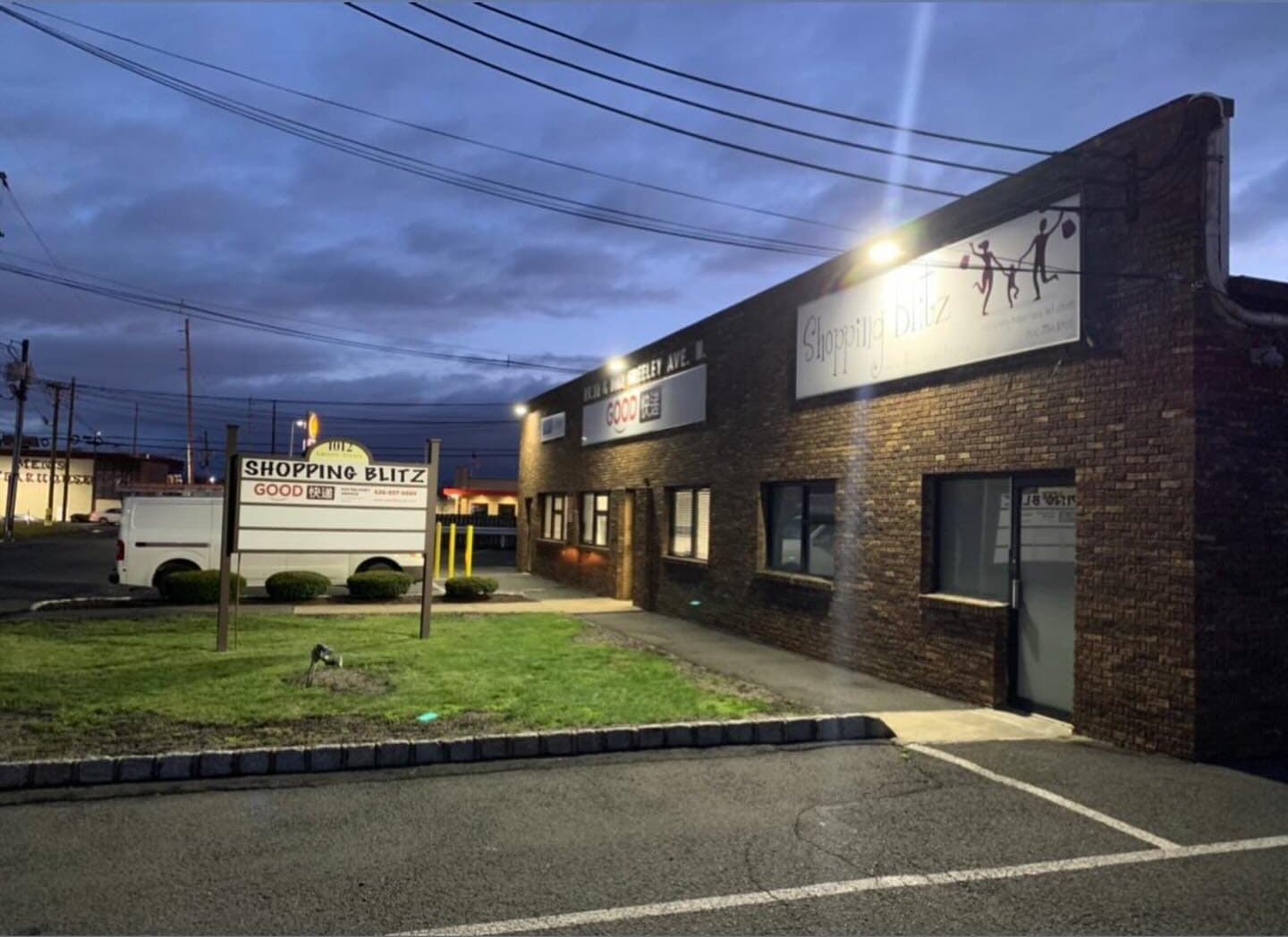 A brick commercial building at dusk with its wall lights on over the parking lot